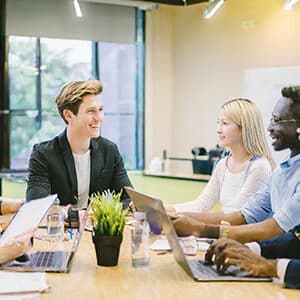 Un groupe multiethnique d’hommes et de femmes en formation, assis autour d’une table dans une salle moderne, échangeant des idées et souriant, illustrant la collaboration et le leadership.