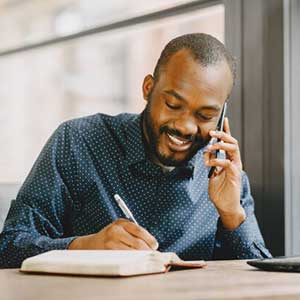 Un homme souriant en plein appel téléphonique, prenant des notes sur un carnet tout en utilisant un ordinateur portable, symbolisant la flexibilité et la productivité professionnelle avec Leaderseed.